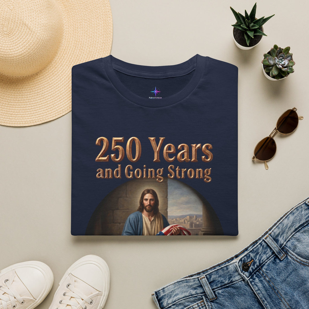 Flat lay view of a folded navy blue patriotic Christian t-shirt with gold text reading '250 Years and Going Strong'. The shirt is styled on a neutral background next to a straw hat, sunglasses, jeans, white sneakers, and small potted plants.